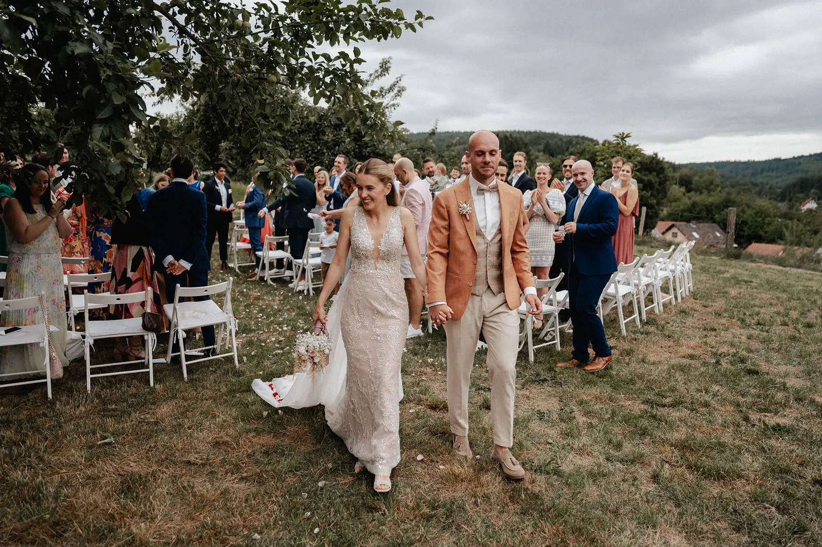 Auszug nach der freien Trauung: Brautpaar geht Hand in Hand durch die Reihen, Gäste applaudieren – Spessarthof, Aura im Sinngrund (bei Mittelsinn, Bayern).