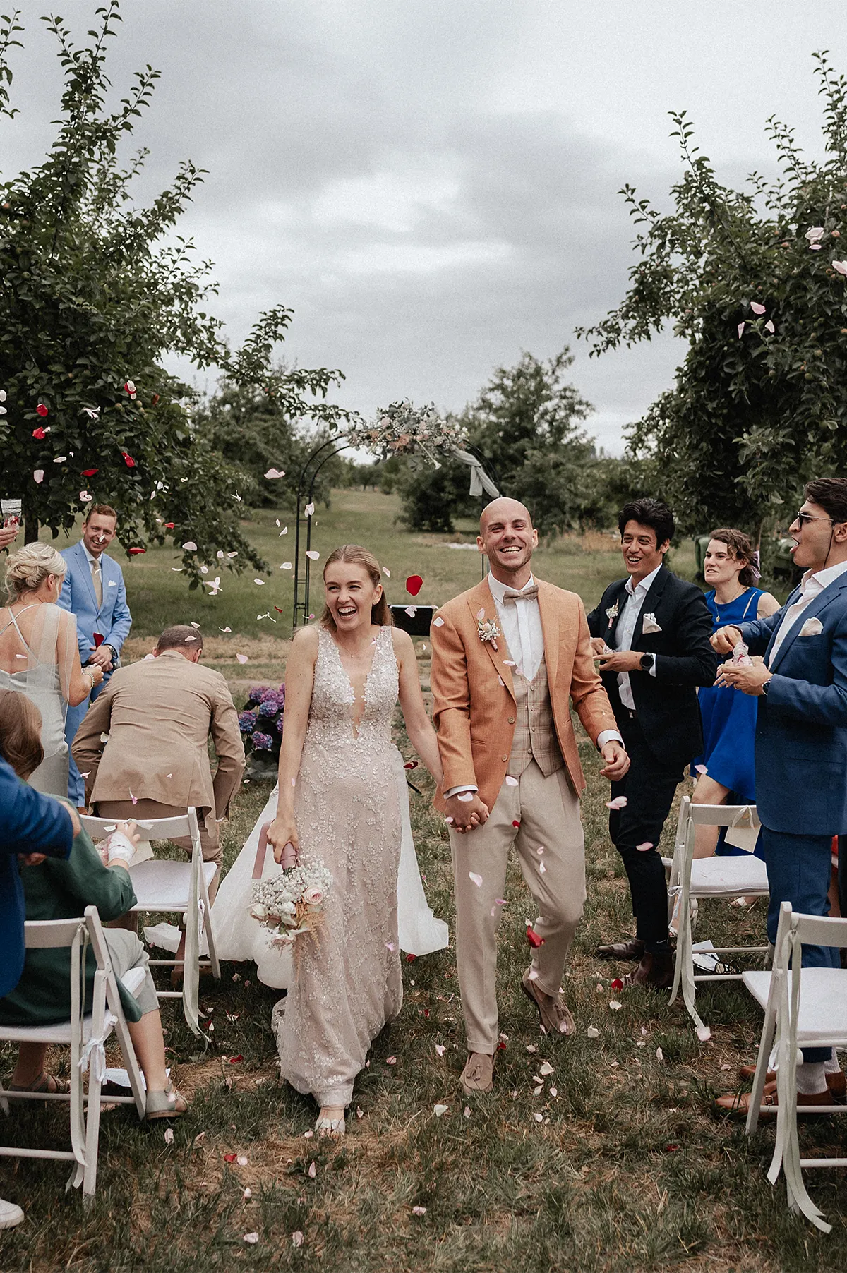 Brautpaar läuft lachend nach der freien Trauung durch den Obstgarten am Spessarthof in Aura im Sinngrund, während Gäste Blütenblätter werfen.