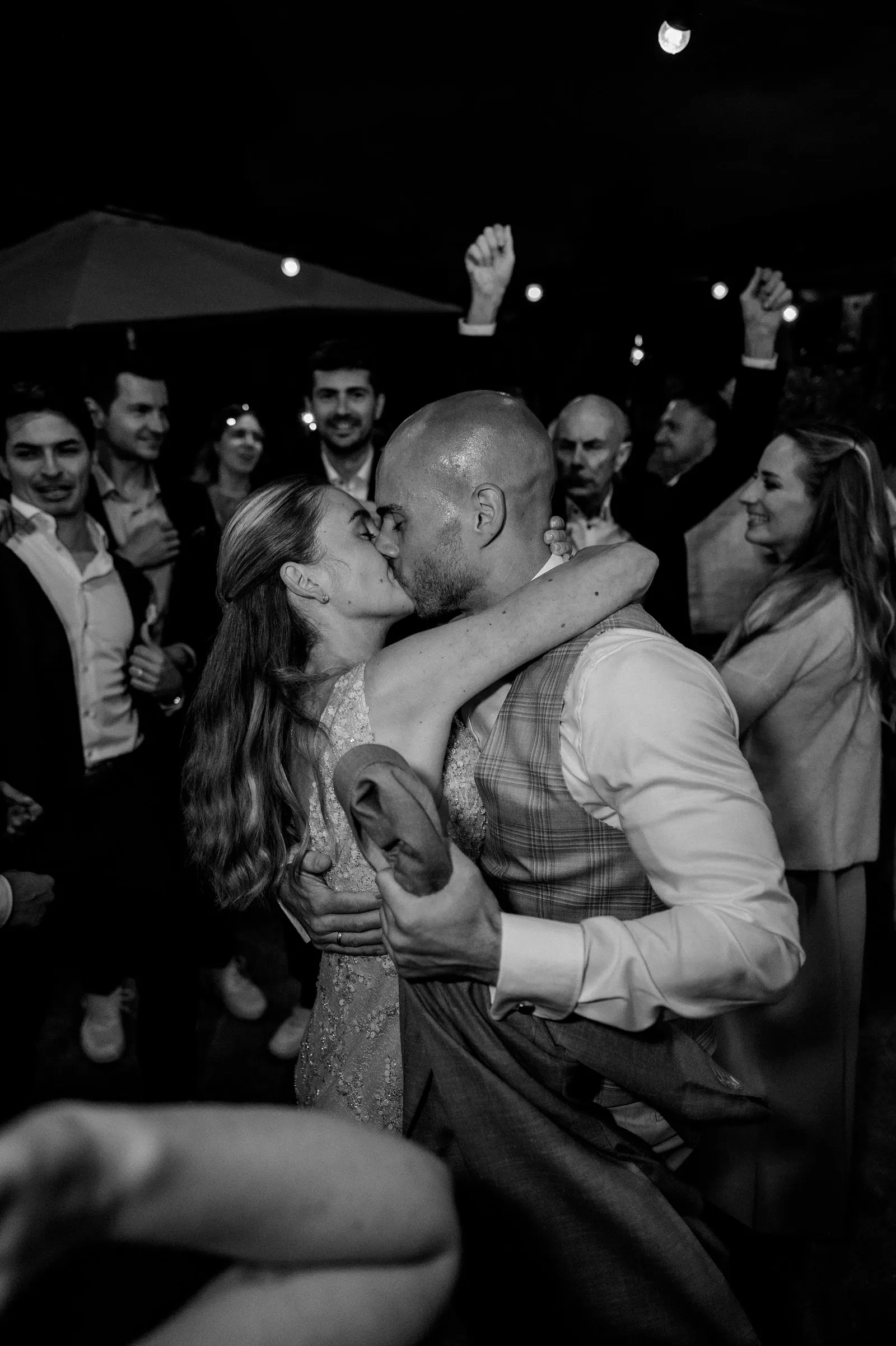 Schwarzweiß-Aufnahme: Das Brautpaar küsst sich eng umarmt auf der Tanzfläche, Gäste jubeln im Hintergrund – Spessarthof, Aura im Sinngrund, Bayern (Landkreis Main-Spessart).