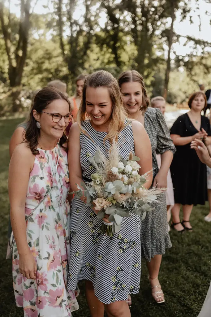 Zwei Freundinnen lachen zusammen, eine hält den Brautstrauß – Moment mit Hochzeitsgästen bei TCA Asfahl in Wuppertal.