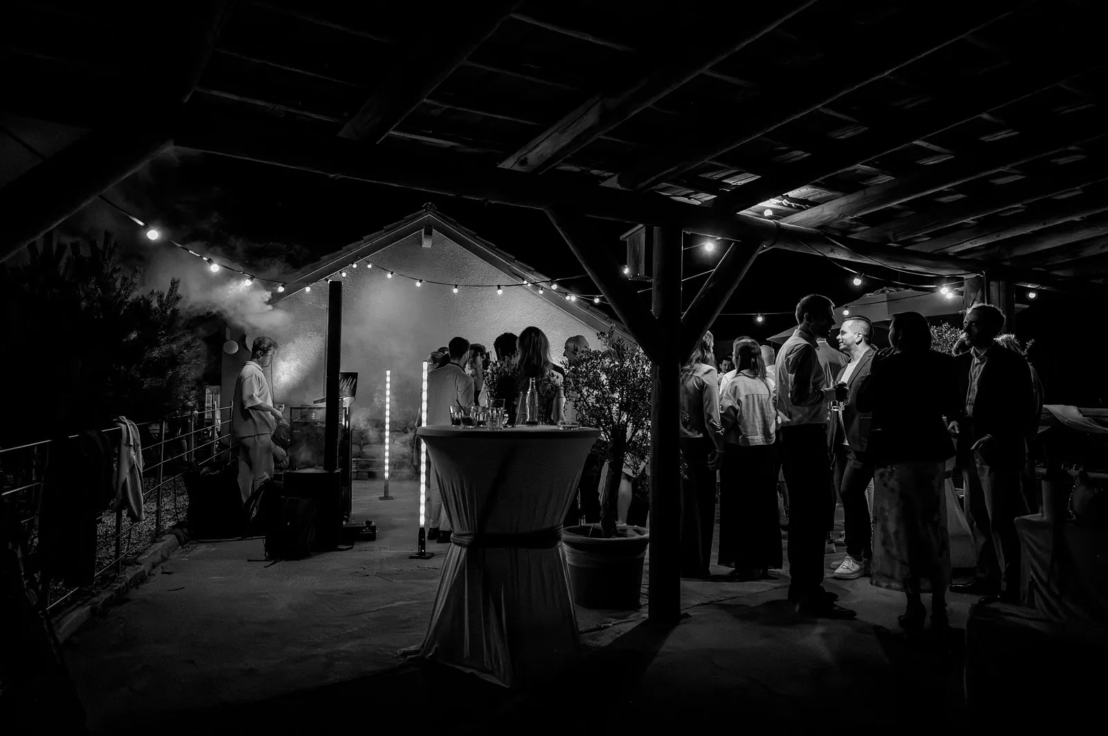 Schwarzweiß-Aufnahme: Gäste stehen abends im Hof unter Lichterketten und einem Holzdach, Stehtische im Vordergrund, leichte Rauchstimmung bei der Feier – Spessarthof, Aura im Sinngrund, Bayern (Unterfranken).