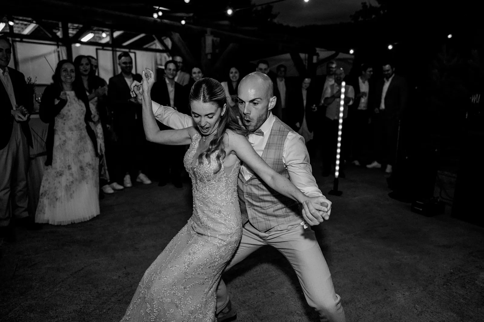 Schwarzweiß-Aufnahme: Brautpaar tanzt ausgelassen auf der Hochzeitsparty, Gäste im Hintergrund – Spessarthof, Aura im Sinngrund, Bayern (Unterfranken).